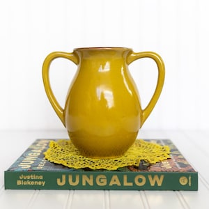May include: A mustard-yellow ceramic pitcher with two handles, sitting on a green book with the word "JUNGALOW" in gold lettering. A yellow lace doily is beneath the pitcher. The pitcher has a glossy finish.
