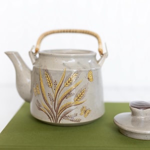 May include: A light gray ceramic teapot with a speckled finish, featuring a golden wheat stalk and butterfly illustration on the side. The teapot has a curved spout and a light-colored woven handle. A matching lid with a dark brown knob sits nearby.