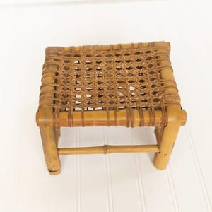 May include: A small, rectangular stool constructed from light brown bamboo poles. The seat features a woven lattice pattern made from thin, dark brown strips of material. The frame includes four sturdy legs connected by horizontal support bars.