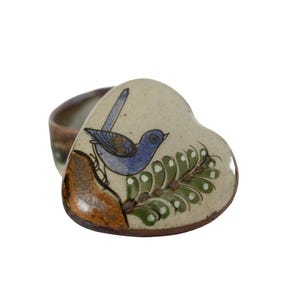 May include: A heart-shaped ceramic trinket box with a cream-colored lid featuring a hand-painted blue bird perched on a brown branch next to a green leafy sprig. The base of the box is a dark brown ceramic with a smooth, glazed finish.