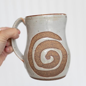 May include: A ceramic mug with a speckled light gray glaze and a large, textured brown spiral design on the front. The mug features a curved handle on the left side and a slightly flared rim, showcasing a handcrafted appearance with visible clay texture.