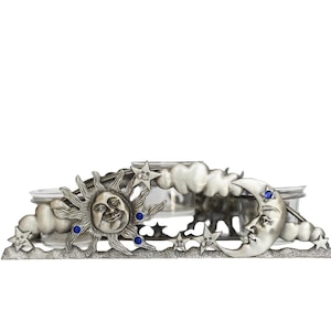 May include: A silver-toned metal candle holder featuring a celestial design with a smiling sun, a crescent moon, and clouds. The piece is accented with small, embedded blue rhinestones and includes clear glass inserts for holding candles.