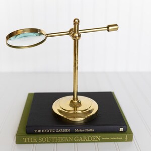 May include: A brass magnifying glass on a stand, resting on two books. The magnifying glass has a circular lens and a long arm. The books are green and black, with the titles "THE EXOTIC GARDEN" and "THE SOUTHERN GARDEN".
