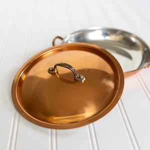 May include: A round copper lid with a central brass handle positioned in front of a shallow, circular copper pan. The pan features a metallic silver-toned interior lining and two small brass side handles attached to the exterior rim.