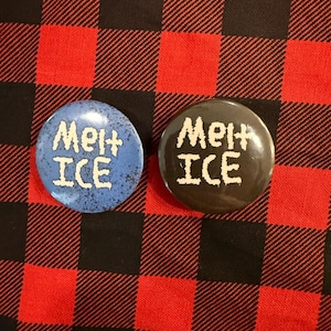 May include: Two round buttons with the text "Melt ICE" in white. One button is blue with white speckles, the other is black. The buttons are on a red and black plaid fabric background.