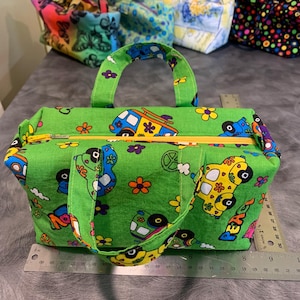 May include: A green fabric tote bag with a yellow zipper and a matching handle. The bag features a colorful print of cartoon vans, flowers, and peace signs. A ruler shows the bag is approximately 9 inches long.