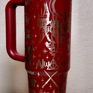 Puede incluir: Un vaso t&eacute;rmico rojo con asa, con un dise&ntilde;o plateado de un castillo, estrellas y texto. El vaso incluye las palabras "Always" y "Catch me if you can". La tapa es blanca y el asa es roja.