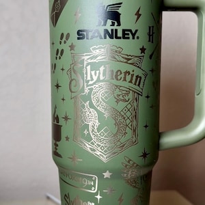 Puede incluir: Vaso Stanley verde con diseños de "Harry Potter" y "Slytherin". El vaso tiene un asa y una tapa negra. El diseño incluye el escudo de Slytherin, rayos y estrellas. También es visible el texto "Ambitious Leadership".