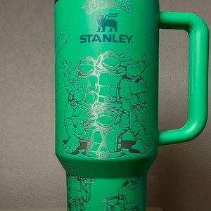 Pu&ograve; includere: Bicchiere Stanley verde con grafica dei Tartarughe Ninja. Il bicchiere ha un coperchio bianco e argento, un manico verde e una cannuccia. Il design include le Tartarughe e il logo Stanley. Questo bicchiere isolato &egrave; ideale per bevande fredde.