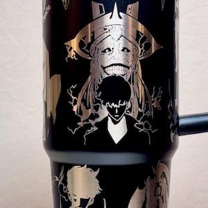 Pode incluir: Caneca preta de aço inoxidável com desenhos prateados gravados. A caneca apresenta ilustrações de personagens de anime. O logotipo da Stanley é visível. A caneca tem uma alça e uma tampa.