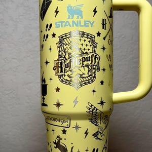 Puede incluir: Un termo Stanley amarillo con gráficos de Harry Potter y Hufflepuff. El termo tiene un asa y tapa amarillas, con varios símbolos y texto, incluyendo "Platform 9 3/4". El termo está decorado con estrellas, rayos y un búho.