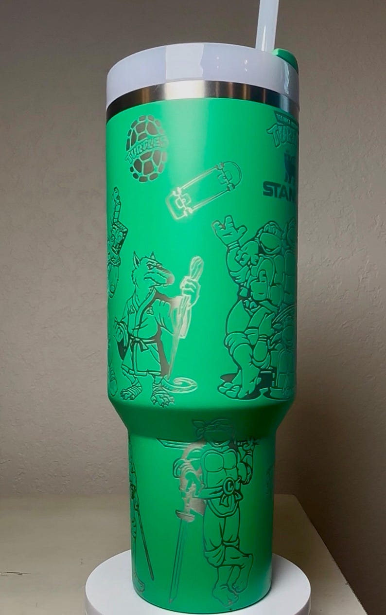Pu&ograve; includere: Un bicchiere in acciaio inossidabile verde con illustrazioni dei personaggi delle Tartarughe Ninja e grafica correlata. Il bicchiere ha un coperchio bianco e una cannuccia. Il design include uno skateboard e la parola "STAN".