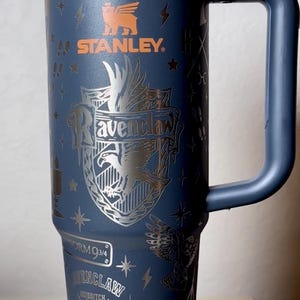 Puede incluir: Un termo de viaje Stanley azul marino con diseños de "Harry Potter" y "Ravenclaw". El termo tiene un asa y una tapa negra. El escudo de Ravenclaw y otros gráficos son plateados. El logotipo de Stanley es naranja.