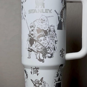 Puede incluir: Taza de viaje Stanley blanca con ilustraciones negras de personajes de anime. La taza tiene un asa grande y una tapa blanca. El logotipo de Stanley es visible. La taza está decorada con texto japonés y personajes de anime.