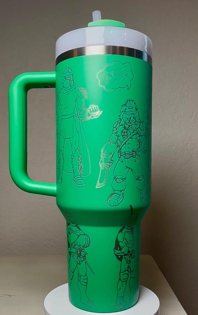 Pu&ograve; includere: Bicchiere termico verde con manico, con illustrazioni incise di personaggi dei cartoni animati e la parola "DUDE" in un fumetto. Il bicchiere ha una parte superiore bianca e una fascia argentata.