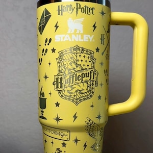 Puede incluir: Vaso Stanley amarillo con gráficos de Harry Potter y Hufflepuff. El vaso incluye el logotipo de Stanley, la plataforma 9 3/4 y otras ilustraciones temáticas. Tiene un asa amarilla y una tapa blanca y amarilla.
