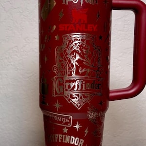 Puede incluir: Un vaso Stanley rojo y dorado con im&aacute;genes de Harry Potter y Gryffindor. El vaso incluye las palabras "Harry Potter", "Gryffindor", "Plataforma 9 3/4" y "Quidditch". Tiene una tapa blanca y roja y un asa roja.