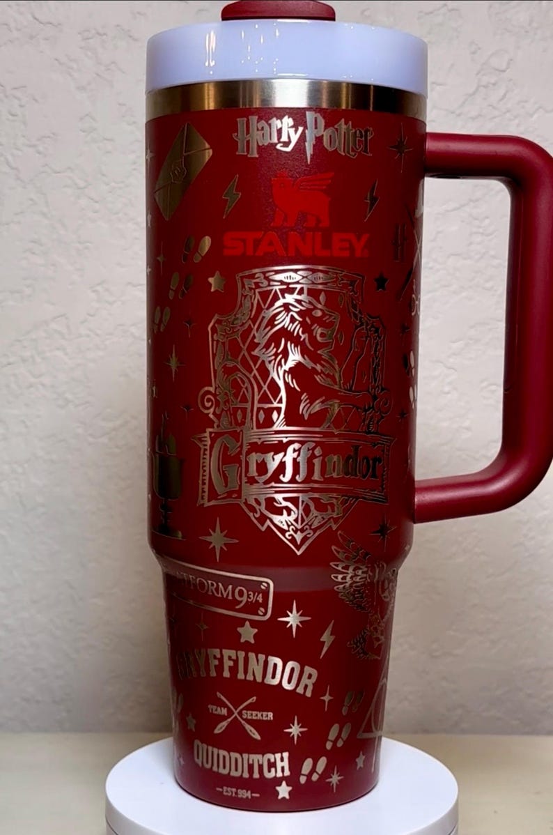 Puede incluir: Un vaso Stanley rojo y dorado con im&aacute;genes de Harry Potter y Gryffindor. El vaso incluye las palabras "Harry Potter", "Gryffindor", "Plataforma 9 3/4" y "Quidditch". Tiene una tapa blanca y roja y un asa roja.