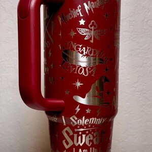 Puede incluir: Un vaso t&eacute;rmico rojo con asa, con dise&ntilde;os plateados grabados y texto de una popular serie de fantas&iacute;a. El vaso incluye frases como "Mischief Managed" y "I Solemnly Swear that I Am Up to No Good."