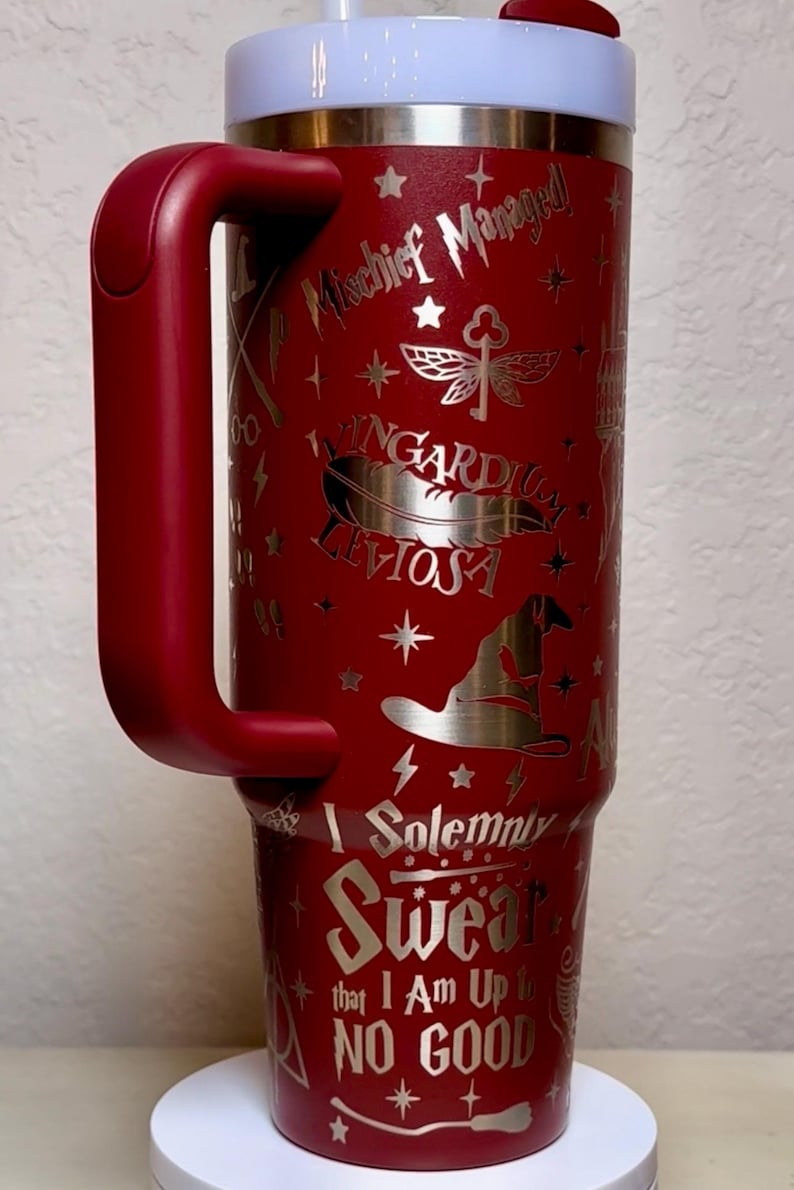 Puede incluir: Un vaso t&eacute;rmico rojo con asa, con dise&ntilde;os plateados grabados y texto de una popular serie de fantas&iacute;a. El vaso incluye frases como "Mischief Managed" y "I Solemnly Swear that I Am Up to No Good."