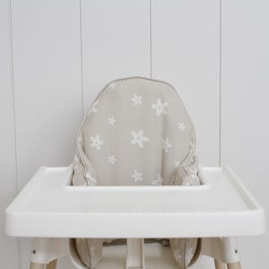 May include: A white high chair with a tray, featuring a beige seat cushion adorned with white floral patterns. The high chair is set against a white paneled wall, creating a clean and simple aesthetic.