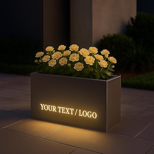 May include: A rectangular, dark grey planter box illuminated from within, displaying the text "YOUR TEXT / LOGO" in a warm, glowing light. The planter is filled with yellow flowers. The scene is set outdoors at night.