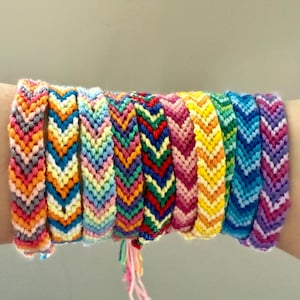 May include: A collection of colorful friendship bracelets, each with a chevron pattern. The bracelets feature a variety of colors, including pink, orange, yellow, green, blue, and purple. The bracelets are handmade and appear to be made of woven thread.