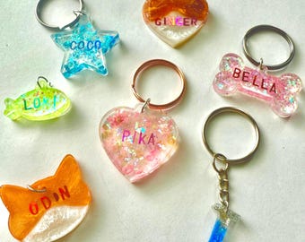 Puede incluir: Una colección de llaveros de resina hechos a mano en varias formas, incluyendo una estrella, un corazón, un pez, un hueso, un gato y una pequeña forma de T. Cada llavero presenta purpurina y un nombre, como "COCO", "GINGER", "BELLA", "ODIN", "LOLI" y "PIKA".