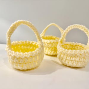 May include: Three small, handmade crochet baskets in a soft yellow hue. Each basket has a rounded base, a cylindrical body, and a curved handle. The baskets are arranged on a white surface, showcasing their delicate texture and design.
