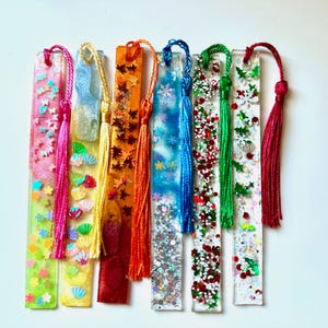 Puede incluir: Una colección de marcadores de resina hechos a mano en varios colores, cada uno adornado con elementos decorativos como purpurina, estrellas y hojas. Cada marcador tiene una borla a juego. Los marcadores miden aproximadamente 15 cm de largo.