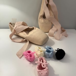 Puede incluir: Un par de zapatillas de ballet con cintas rosas y una colección de pequeñas zapatillas de ballet de ganchillo en rosa, amarillo, azul y negro. Los zapatos más grandes son de color beige claro.