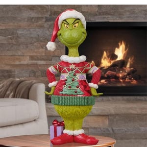 Könnte beinhalten: Eine grüne Charakterfigur mit einer rot-weißen Weihnachtsmannmütze, einem roten Pullover mit grünem Weihnachtsbaummuster und roten Schuhen. Die Figur steht mit den Händen in den Hüften, hat eine strukturierte Oberfläche und ein kleines lila Geschenk.