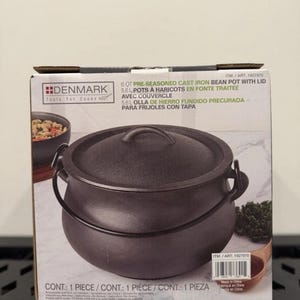 May include: A Denmark Tools For Cooks 5.6 L pre-seasoned cast iron bean pot with lid. The packaging features a black cast iron pot with a rounded body, two side handles, and a matching lid. The box displays the product name and a barcode.
