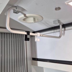 Może przedstawiać: Srebrny, zakrzywiony metalowy system nośny, zamontowany do białego sufitu. System obejmuje poziomy drążek z czarnymi i białymi plastikowymi klipsami. Projekt wydaje się funkcjonalny i nowoczesny, odpowiedni do zastosowań wewnętrznych.