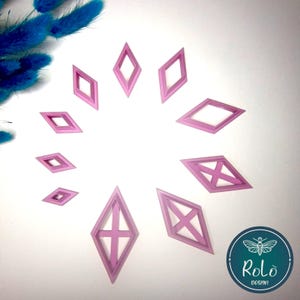 Può includere: Una collezione di oggetti rosa a forma di diamante disposti in un motivo circolare. Alcuni diamanti hanno disegni a croce o a X all'interno. Gli oggetti sono su una superficie bianca, con un gruppo di elementi decorativi blu nell'angolo in alto a sinistra.