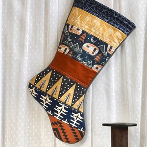May include: A handmade Christmas stocking with a camping theme. The stocking features a patchwork design with various patterns, including camper vans, trees, and geometric shapes in navy, rust, and gold. The stocking is hanging against a white curtain.