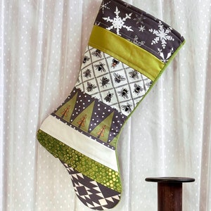 May include: A handmade Christmas stocking with a patchwork design. The stocking features various patterns, including bees, snowflakes, and trees, in shades of green, white, and gray. A vintage iron and wooden spools are in the foreground.
