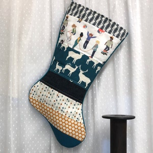 May include: A handmade Christmas stocking with a teal base and various patterned fabric panels. The stocking features a white fabric panel with illustrations of children playing, and a teal panel with white deer silhouettes. A black and white patterned cuff.