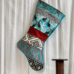 May include: A handmade Christmas stocking with a teal, burgundy, and patterned design. The stocking features a variety of patterns, including rabbits, snowflakes, and geometric shapes. The stocking is hanging against a white curtain.