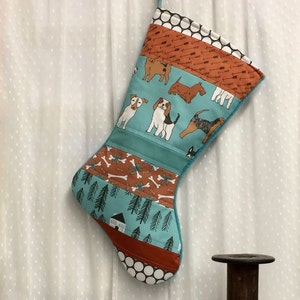 May include: A quilted Christmas stocking with a teal background featuring various dog breeds. The stocking has sections with bone and arrow patterns, and a white polka dot cuff. A teal hanging loop is at the top.