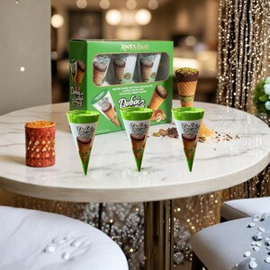 May include: A green box of "Awesome" Dubai ice cream cones with milk chocolate, pistachio cream filling, and a waffle cone. Several cones are displayed on a white marble table, with a small orange candle holder.