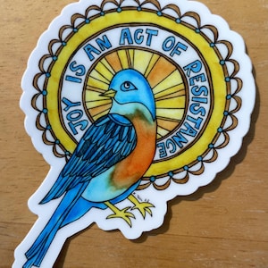 May include: A sticker featuring a bluebird with orange accents, perched in front of a yellow and orange circular design. The text "JOY IS AN ACT OF RESISTANCE" is written around the circle. The sticker has a white border.