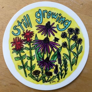 May include: Circular sticker featuring a watercolour illustration of flowers in red, purple, and black, set against a yellow backdrop. The words "still growing" are in teal. The sticker has a white border.