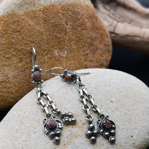 May include: A pair of silver drop earrings featuring small, round, reddish-orange stones set in ornate metalwork. Each earring has a hook closure, a central decorative element, and two dangling chains that end in a detailed, textured silver charm.