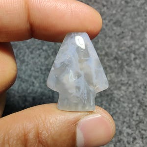May include: A translucent, arrowhead-shaped stone with a light blue and white marbled pattern. The stone is held between fingers, showcasing its polished surface and unique design. The arrowhead has a small, rectangular base.