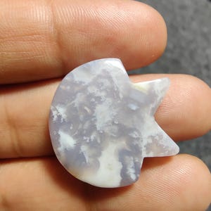 May include: A polished, pale purple gemstone in the shape of a crescent moon with a star-like point. The stone features a cloudy, white pattern. The gemstone is held in a hand.