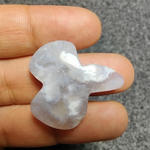 May include: A light blue and white agate stone in an unusual shape. The stone has a smooth, polished surface with a cloudy pattern. The stone is held in a hand against a grey background. The stone is approximately 5 cm long.