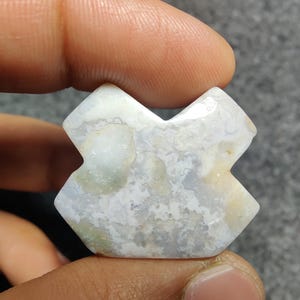 May include: A cross-shaped gemstone in shades of white and grey is held between fingers. The stone features a marbled pattern with hints of light green and beige. The edges are faceted, and the shape is symmetrical.