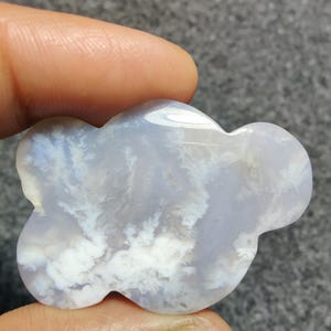 May include: A cloud-shaped gemstone, polished to a smooth finish, displaying a pale blue and white pattern. The stone is held between two fingers. The white patterns resemble clouds or wisps.