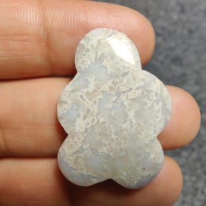 May include: A pale grey, flower-shaped gemstone with a mottled pattern of white and beige. The stone is held in a hand, highlighting its unique shape and natural colour variations. The gemstone is likely used for jewellery or decorative purposes.